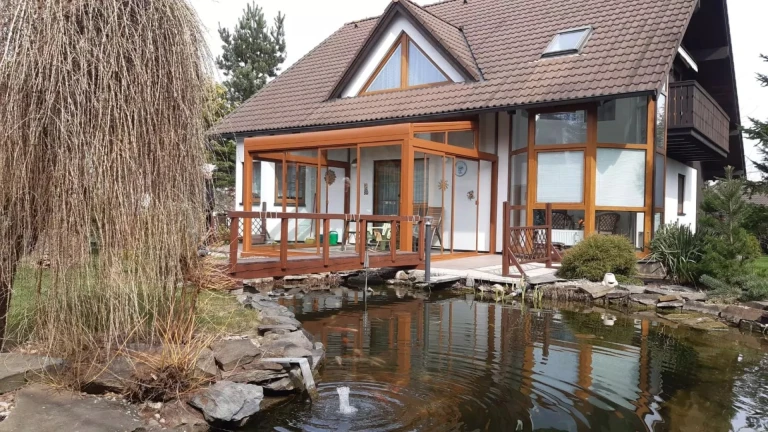 Presklená hliníková pergola pri rodinnom dome so záhradným jazierkom a vŕbou.