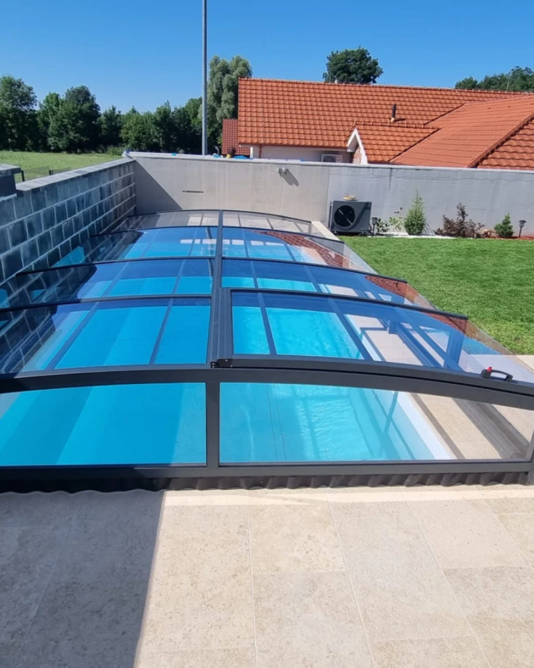 Moderné nízke bazénové prekrytie s čiernym rámom a priehľadným výplňom na záhrade rodinného domu.