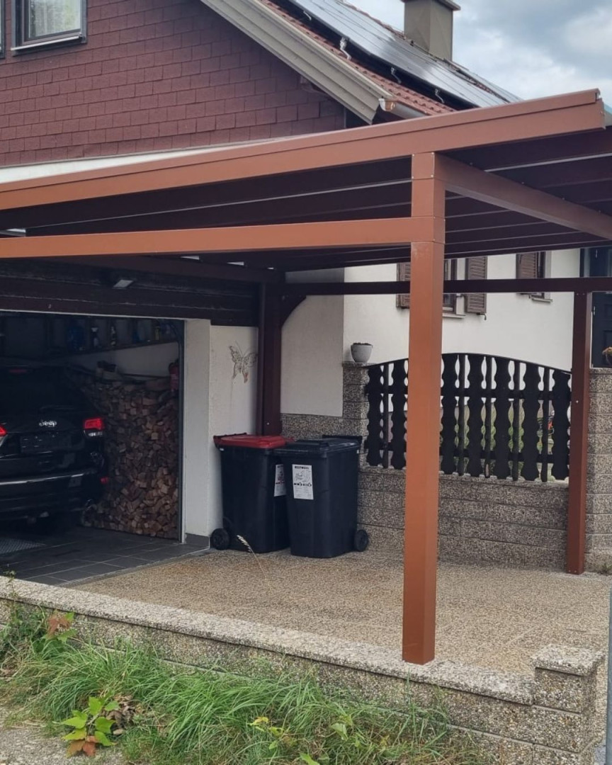 Hnedý prístrešok pre auto vedľa rodinného domu s garážou a smetnými košmi