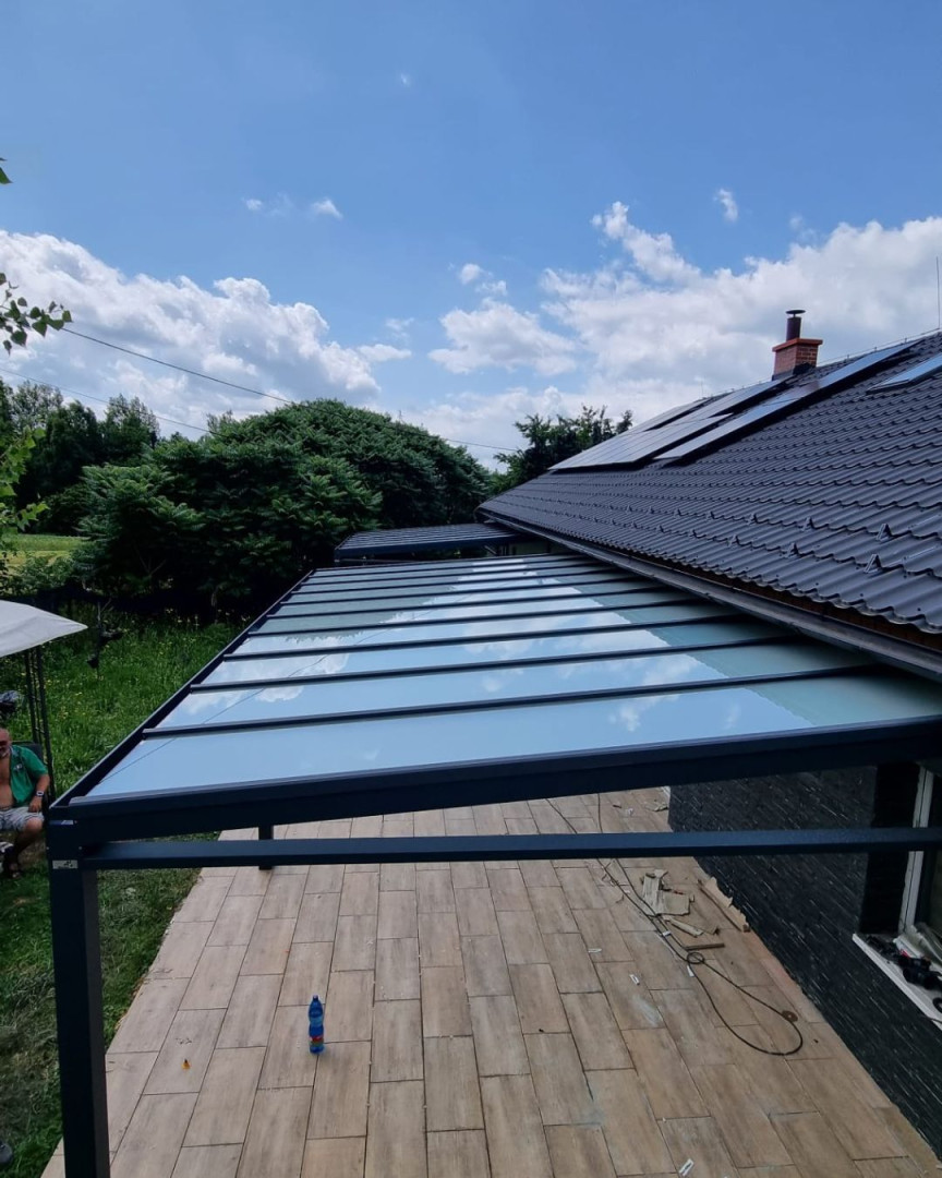 Moderná hliníková pergola so sklenenou strechou pripojená k rodinnému domu s výhľadom do záhrady.