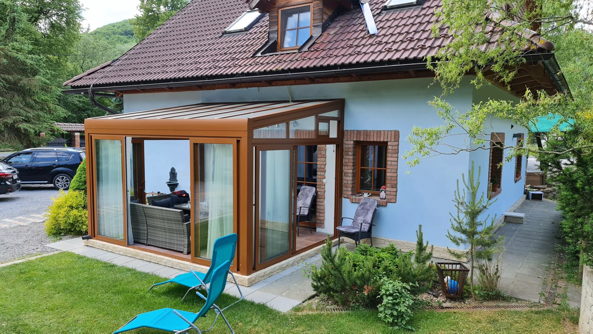 Presklená hliníková pergola ROBUST hnedej farby pristavená k rodinnému domu so záhradným nábytkom.