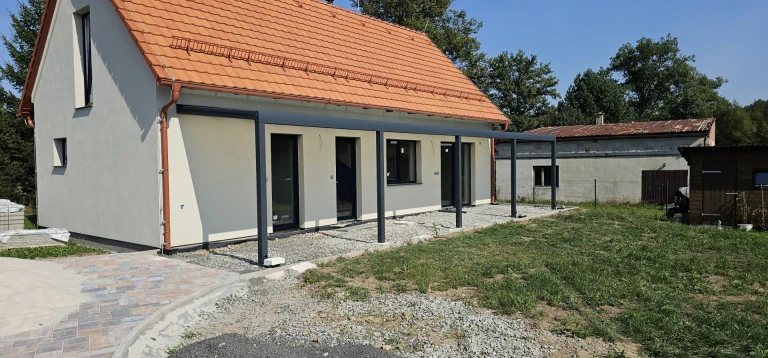 Moderná tmavosivá hliníková konštrukcia pergoly pristavená k novostavbe rodinného domu s oranžovou strechou.