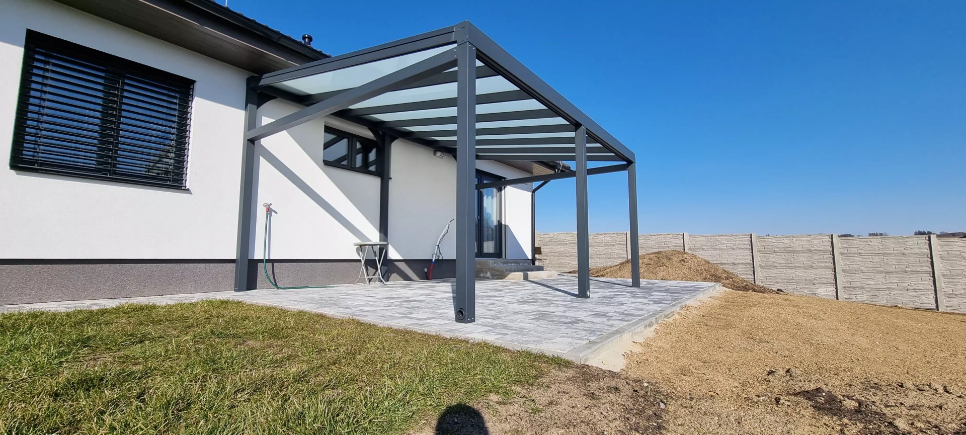Moderná antracitová hliníková pergola so sklenenou strechou pristavená k rodinnému domu na dlaždicovej terase.