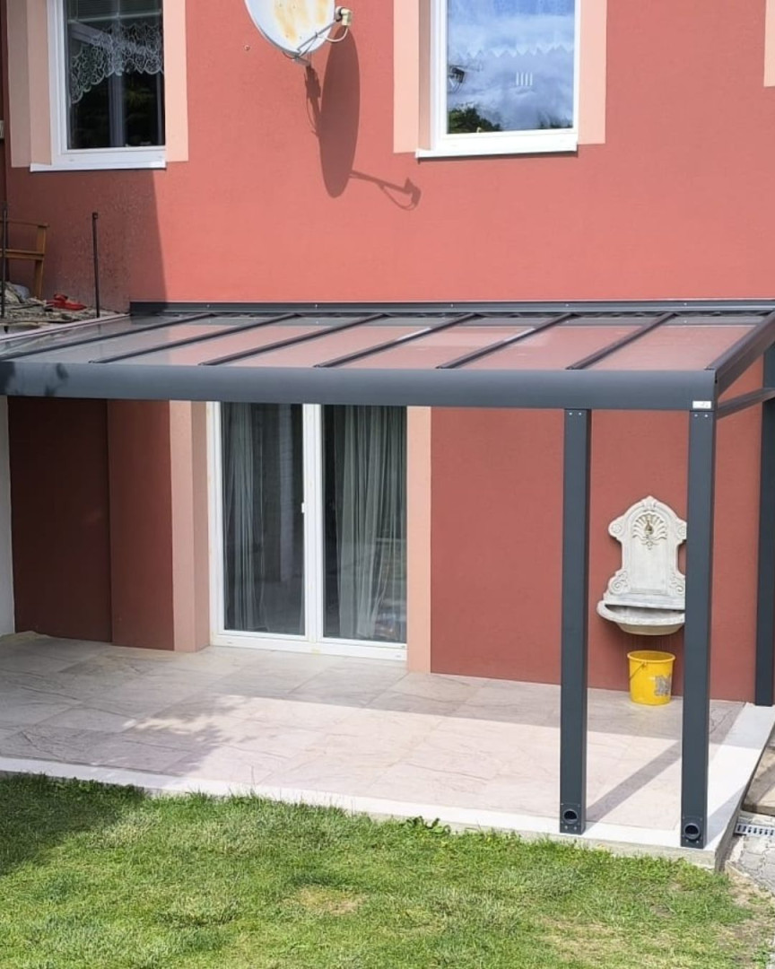 Moderná hliníková pergola s presklenou strechou namontovaná k stene červeného domu nad terasou.