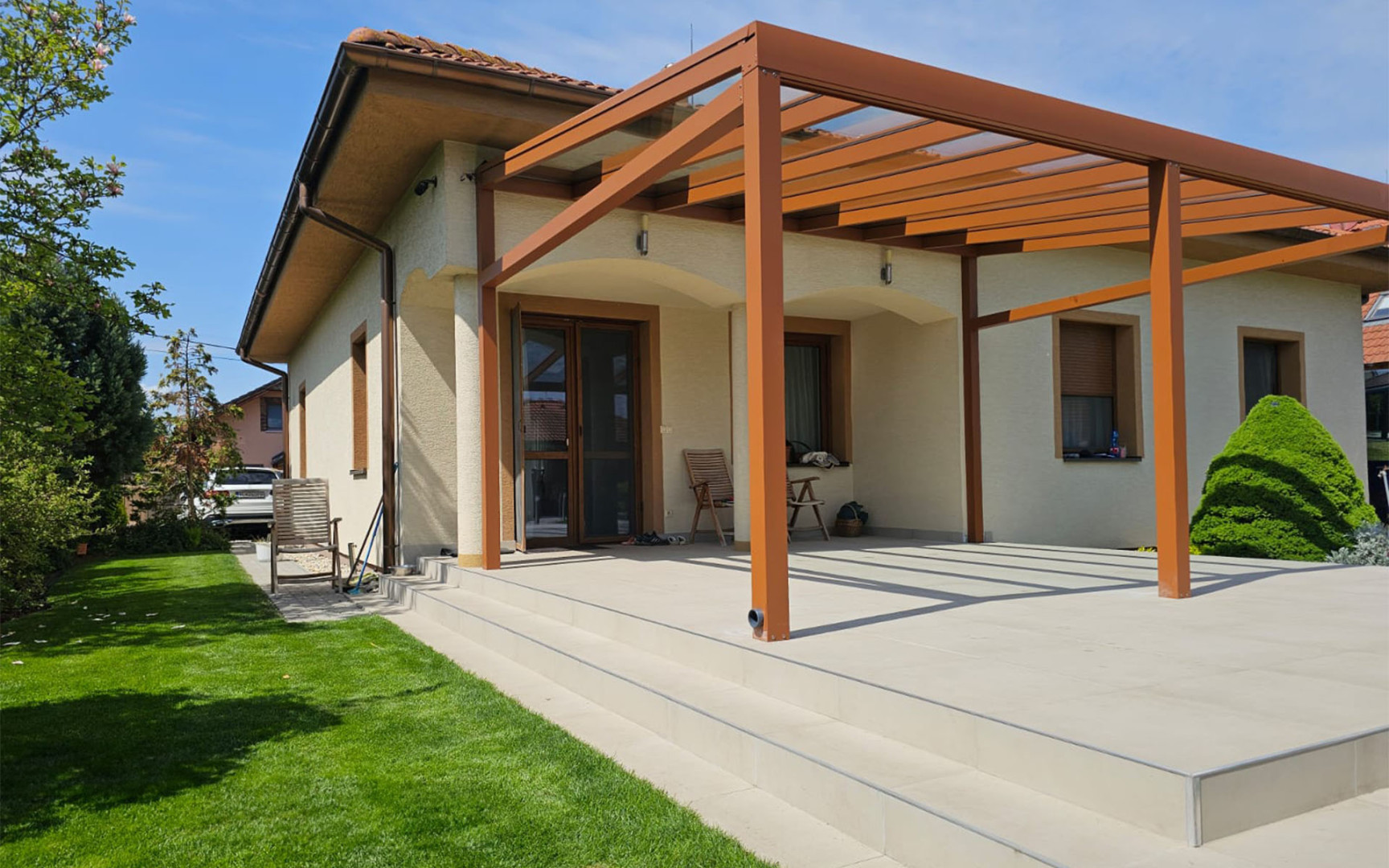 Moderná hliníková pergola v hnedej farbe s presklenou strechou pripojená k rodinnému domu s priestrannou terasou.
