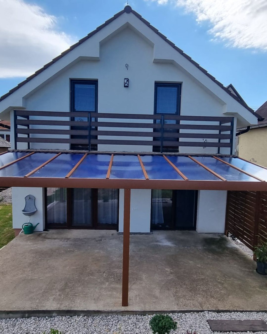 Hnedá hliníková pergola so sklenenou strechou pristavená k rodinnému domu s balkónom.
