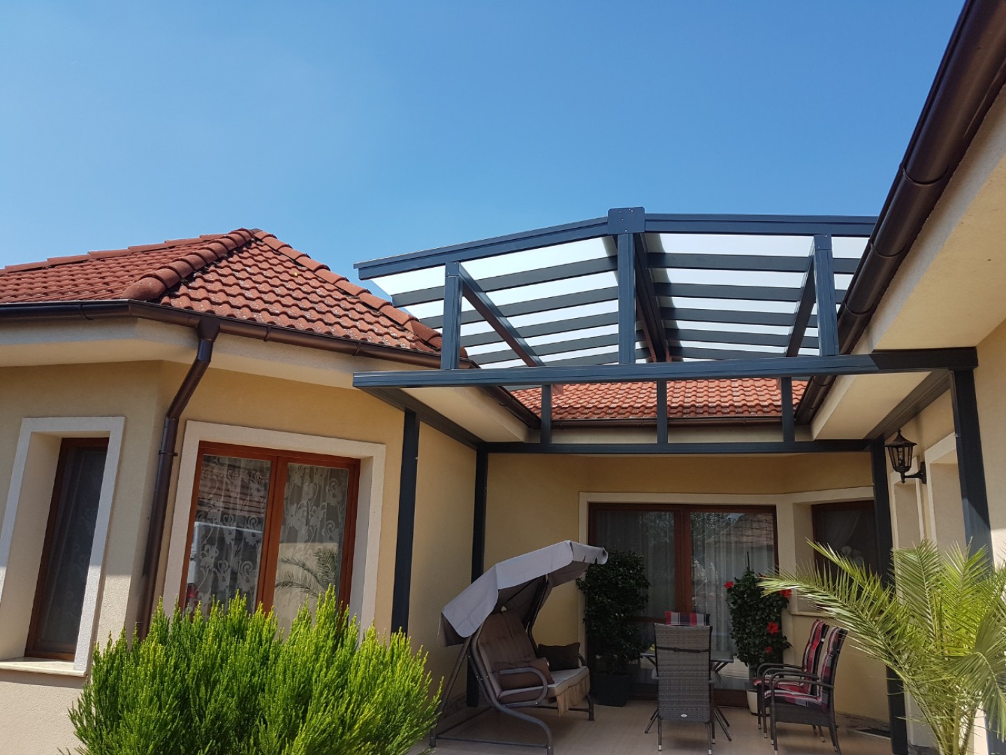 Moderná hliníková pergola ROBUST so zasklenou strechou pripojená k rodinnému domu s terasovým nábytkom a hojdačkou.