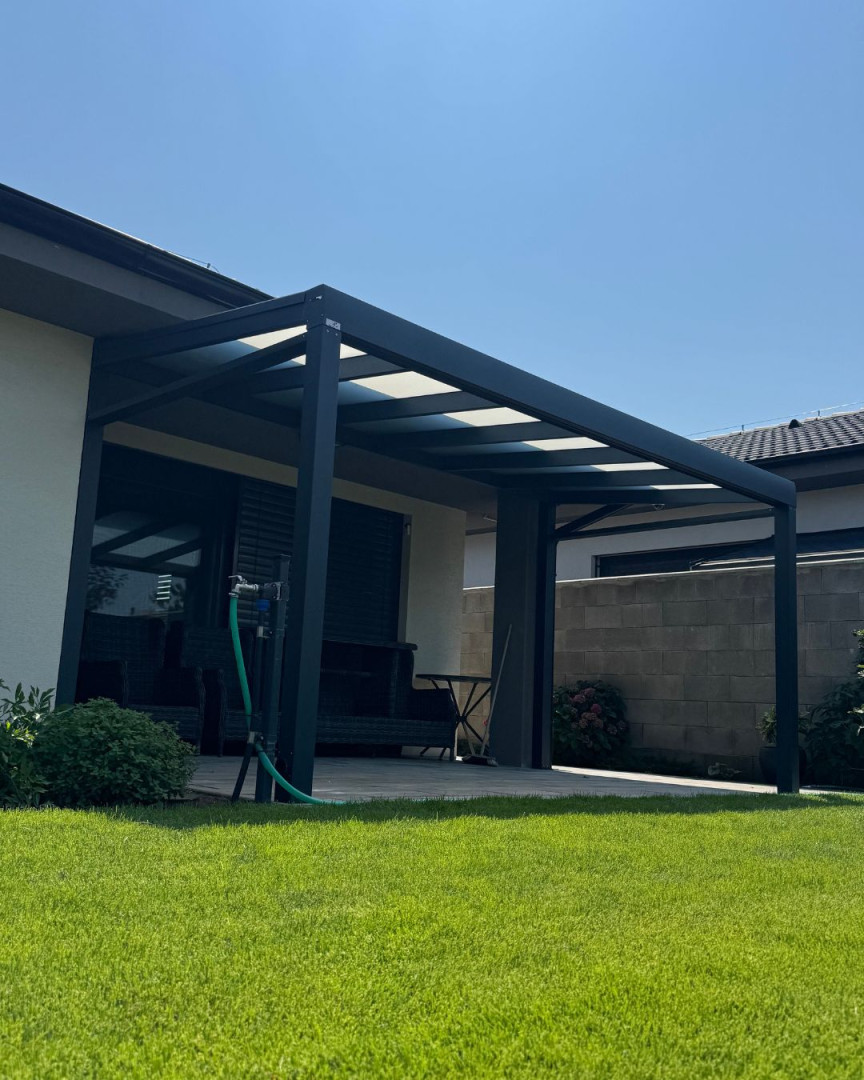 Moderná tmavosivá hliníková pergola pripojená k domu so sklenenou strechou a upraveným trávnikom