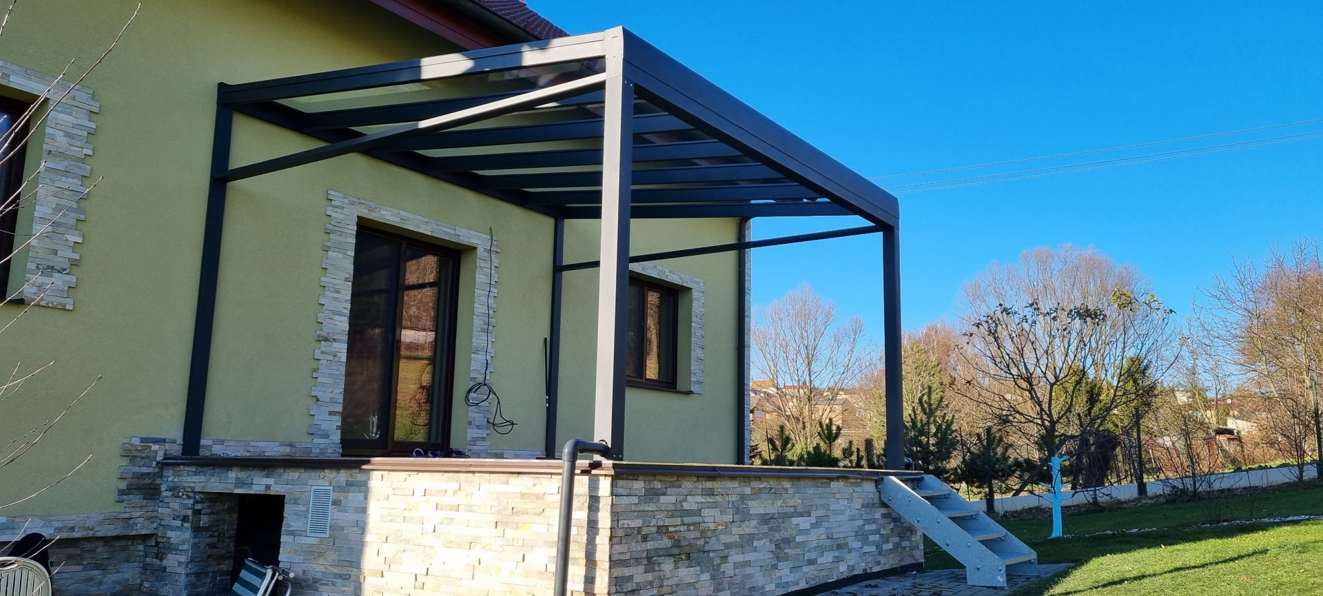 Moderná tmavosivá hliníková pergola ROBUST so sklenenou strechou namontovaná na terase rodinného domu.