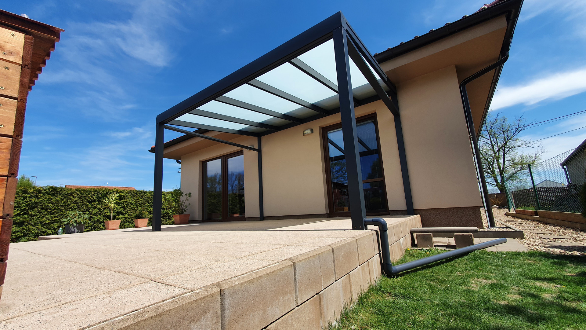 Moderná tmavosivá hliníková pergola so sklenenou strechou namontovaná k rodinnému domu na dláždenej terase.