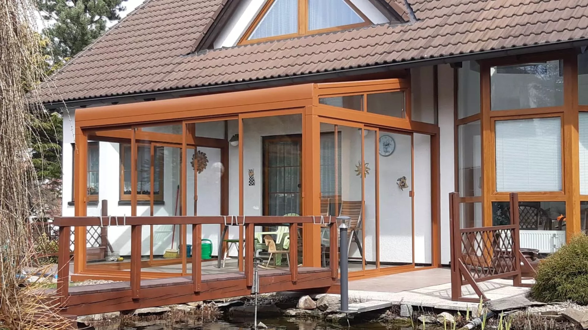 Presklená hliníková pergola ROBUST v hnedej farbe pri rodinnom dome s dreveným mostíkom