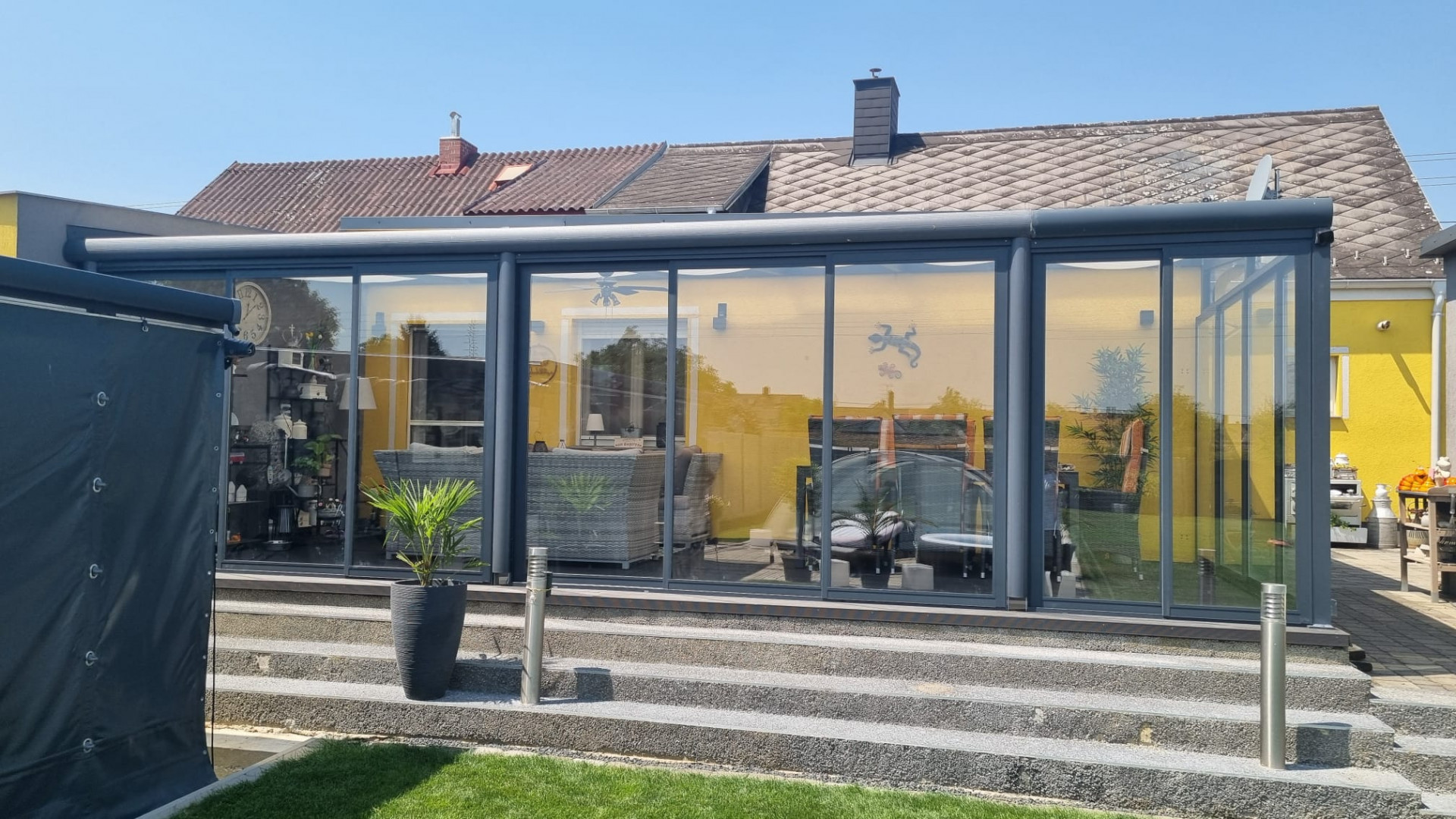 Moderná presklená hliníková zimná záhrada v antracitovej farbe pristavená k žltému rodinnému domu so schodiskom.