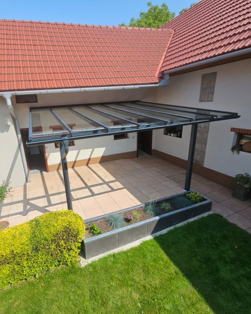 Moderná hliníková pergola so sklenenou strechou pripevnená k stene domu s dláždenou terasou.