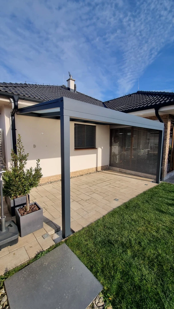 Moderná antracitová hliníková pergola pripojená k domu na dláždenej terase s trávnikom.