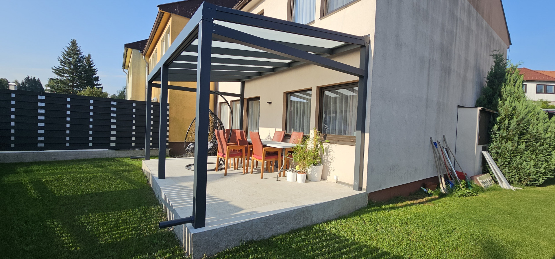 Moderná hliníková pergola s presklenou strechou na betónovej terase rodinného domu so záhradným nábytkom.