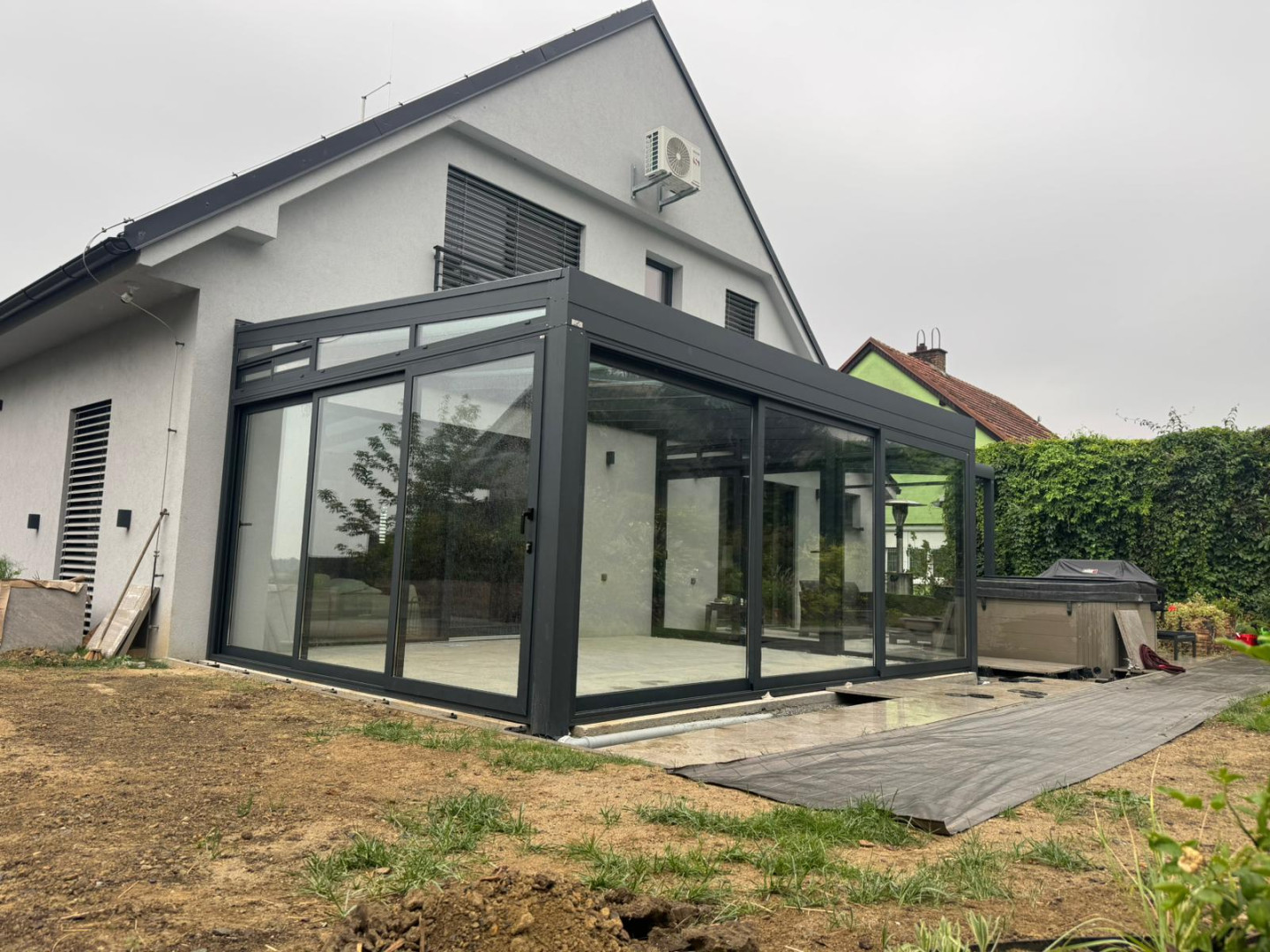 Moderná presklená konštrukcia pripojená k rodinnému domu s tmavým rámom a posuvnými dverami.