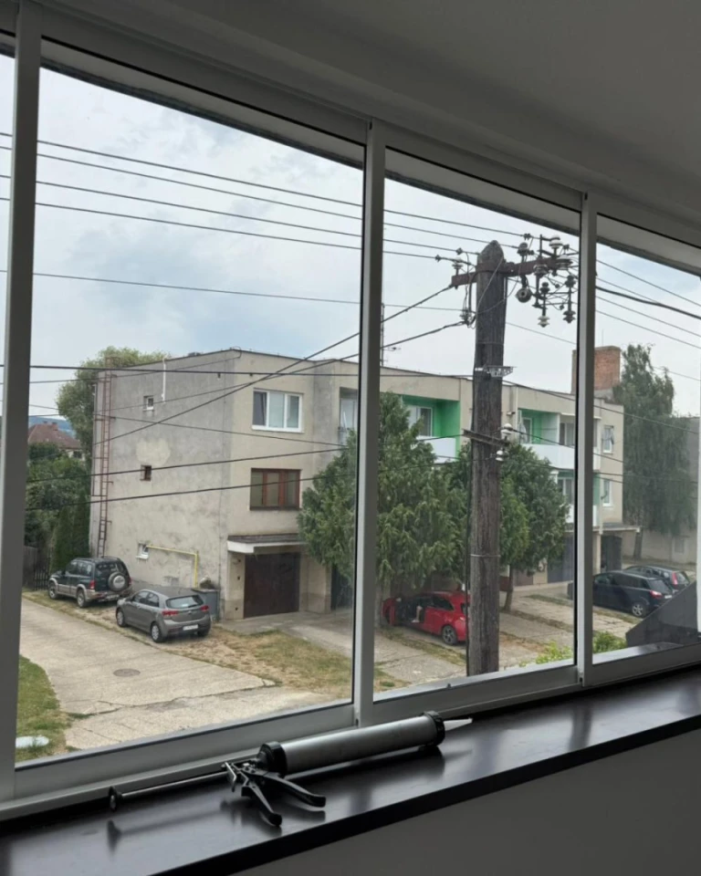 Pohľad cez nové zasklenie na ulicu s bytovými domami a autami, na parapete leží vytlačovacia pištoľ