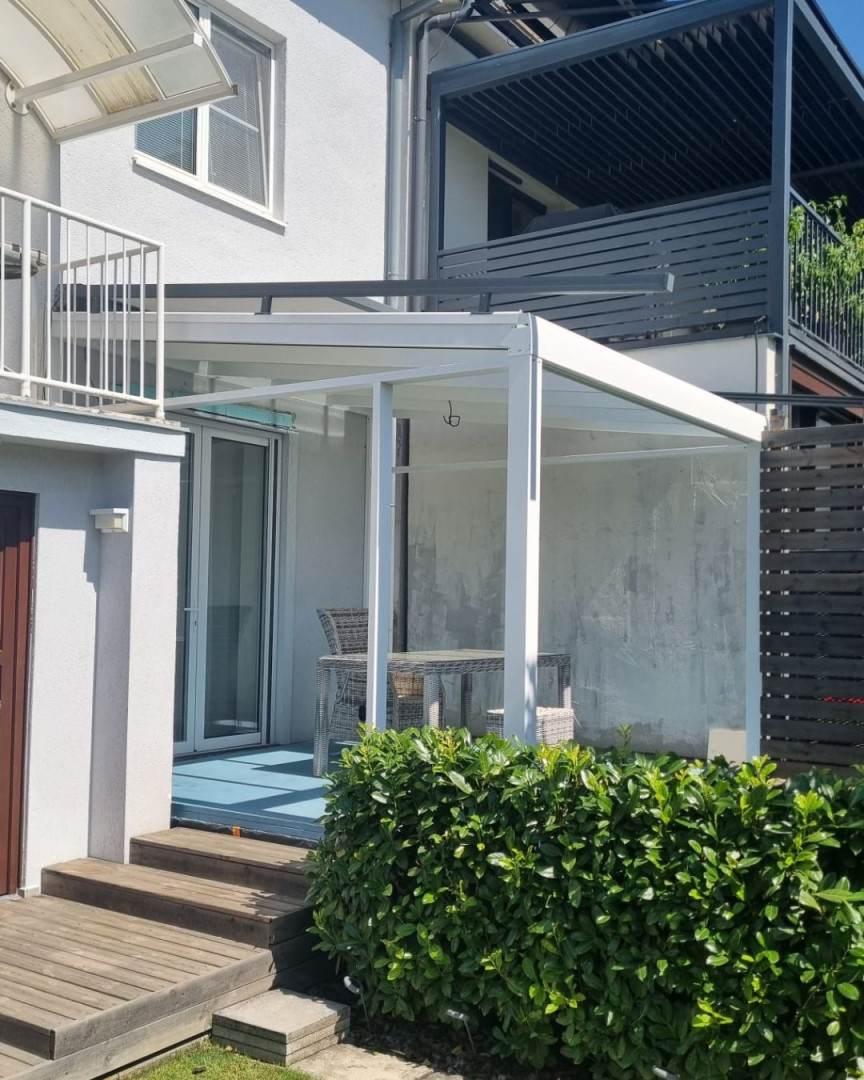 Biela moderná hliníková pergola so sklenenou strechou pri rodinnom dome s terasovým nábytkom.
