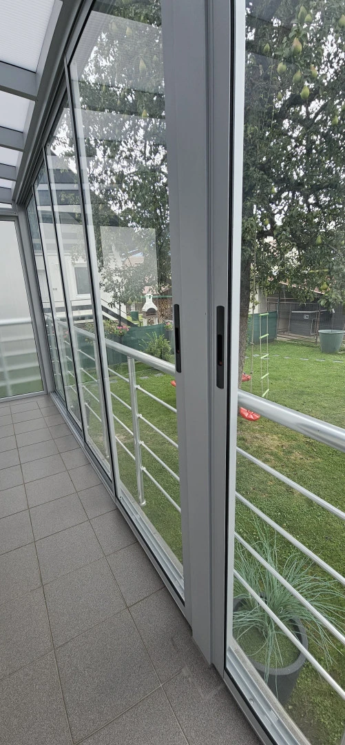 Detailný pohľad na posuvné sklenené dvere a zábradlie zimnej záhrady s výhľadom do zelenej záhrady.
