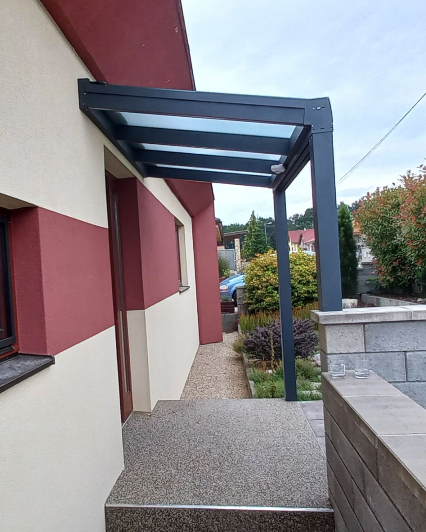 Moderná tmavosivá hliníková pergola so sklenenou strechou namontovaná nad vchodom domu.