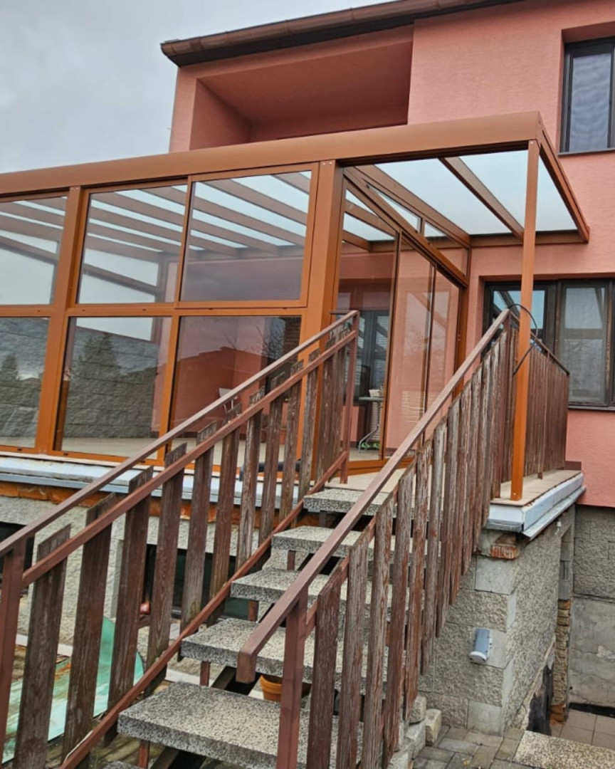 Presklená hliníková pergola hnedej farby namontovaná na terase domu so schodiskom
