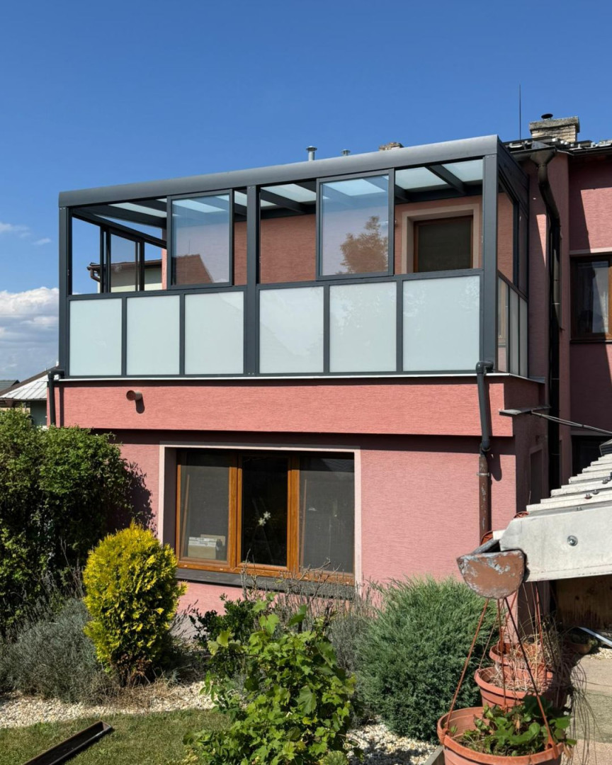 Moderná presklená pergola s tmavošedým hliníkovým rámom a mliečnym sklom na balkóne rodinného domu.